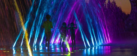 Three children run through an arched manifold of colorful LED ground sprays at night