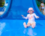 Young girl with sun cap on blue landing pad coming off blue aqua play slide.