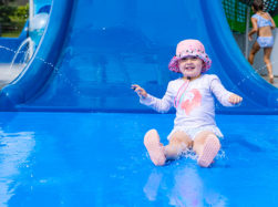 Young girl with sun cap on blue landing pad coming off blue aqua play slide.