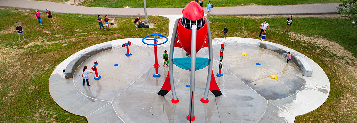 A red and blue space themed splash pad with a central red rocket ship . Various sprayers and water elements surround the central ship along with cement benches.