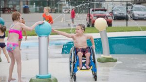 two children play with a spherical squirting water feature on a splash pad. One child is in a wheelchair.