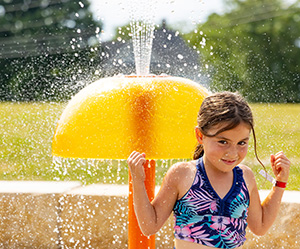 girl standing on front of a yellow spraying aqua dome on a splash pad