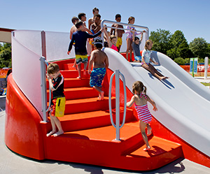 Children climbing to the top of a bright red and white water slide at a spray park