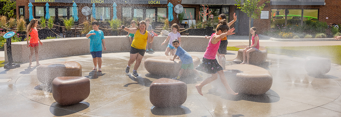 Children running with hands up amid misting ground sprays at Blue Ash Towne Square