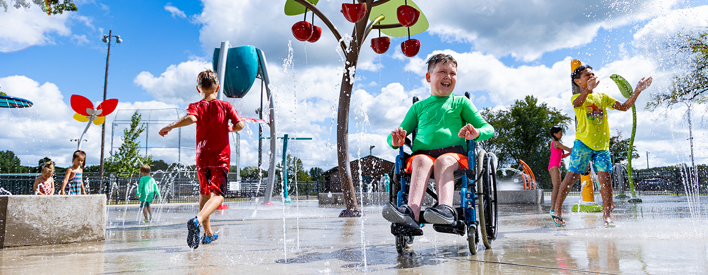 Children playing on a splash pad with a bucket canopy feature in the center and a boy in a wheelchair in the foreground