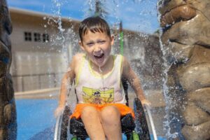 A boy in a wheelchair under a waterfall feature at a splash park