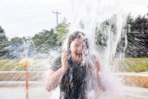 A girl screams with laughter as an overhead bucket dumps water on her at a splash pad